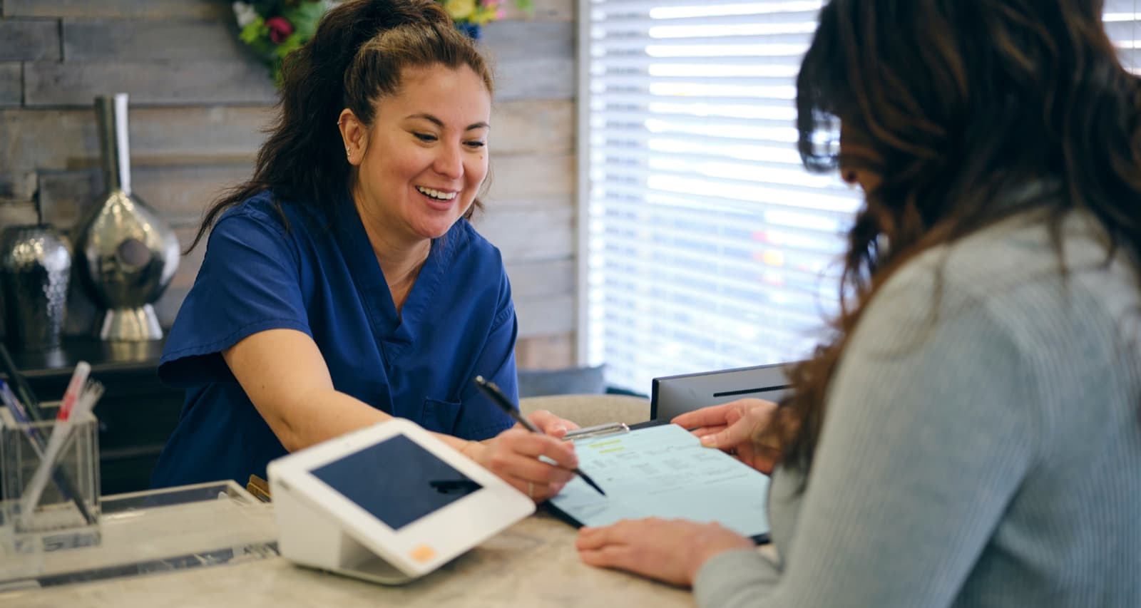 A clinic receptionist in scrubs helping a patient at the front desk